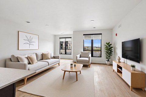 A living room with a grey couch, a wooden coffee table, a flat screen TV, and a large window with a view of the outside.