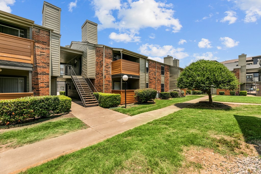 a grassy area with a tree and bushes in front of an apartment building at The Hills at Ironhorse, North Richland Hills
