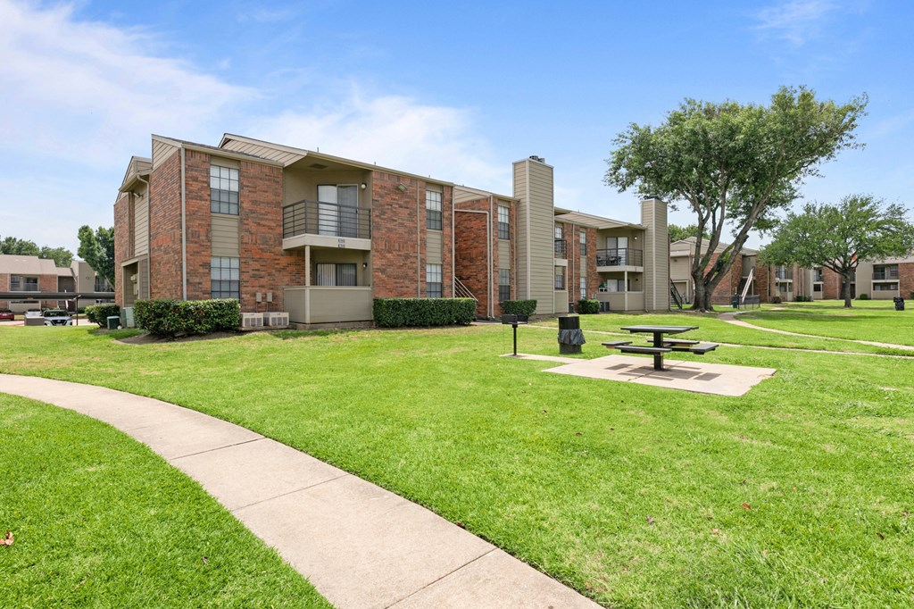 a picnic table sits in the middle of a grassy area in front of an apartment building at The Hudson, North Richland Hills, 76180