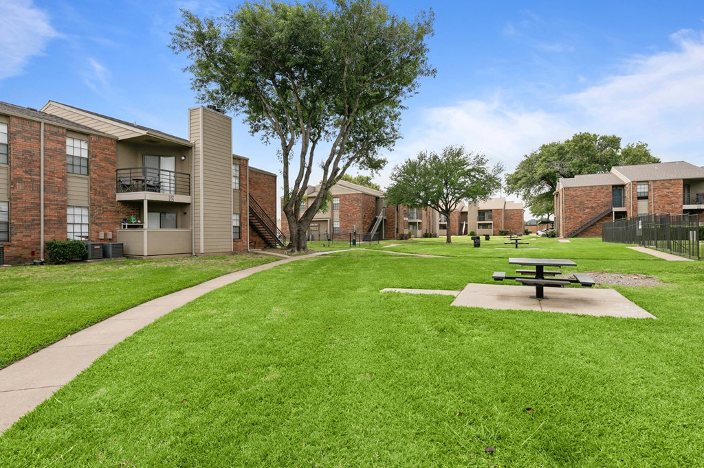 a picnic table sits in the middle of a grassy area in front of a brick building at The Hudson, North Richland Hills, TX