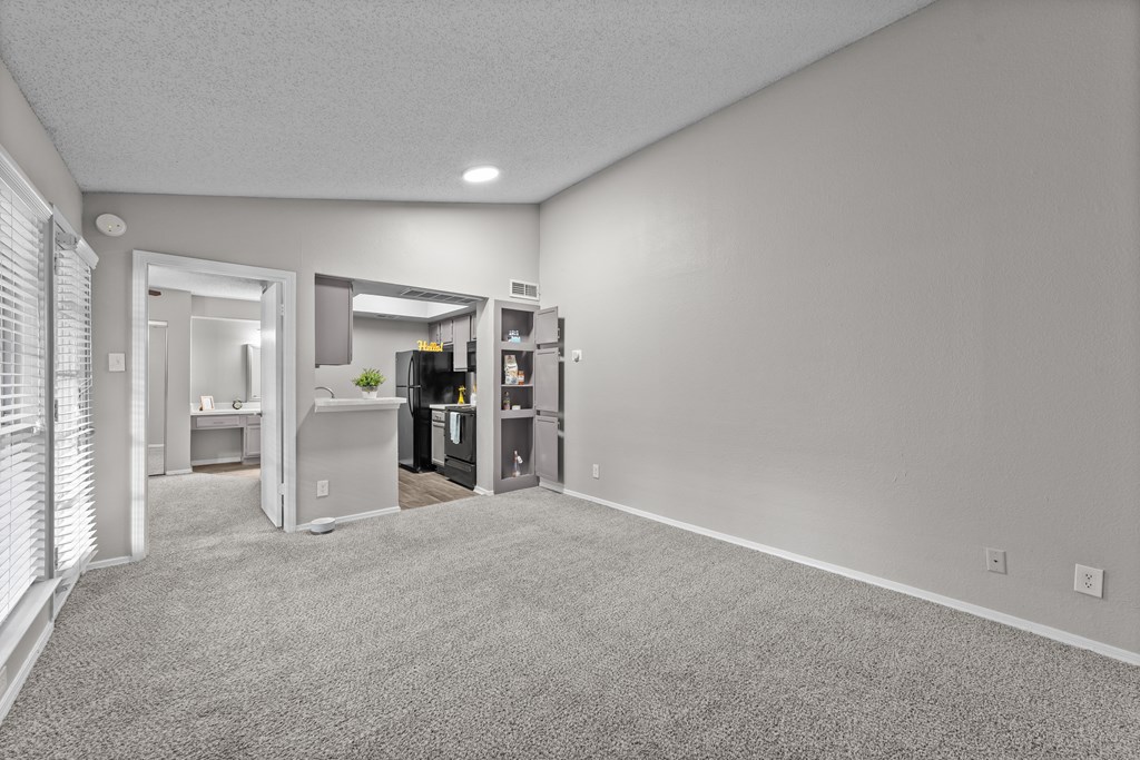 A spacious room with a carpeted floor and a kitchen area visible through the open door.