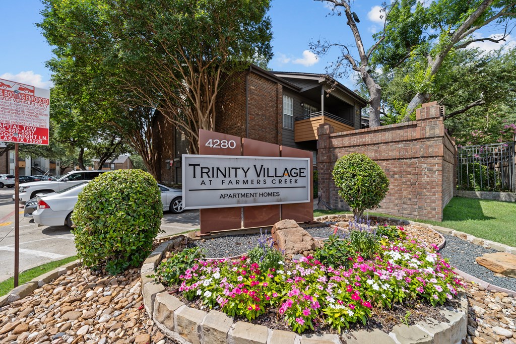A sign for Trinity Village Apartment Homes is surrounded by flowers and greenery.