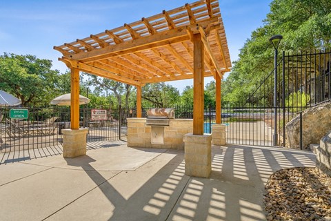 A wooden pergola structure is situated in a park-like setting with a black fence and a stone pillar.