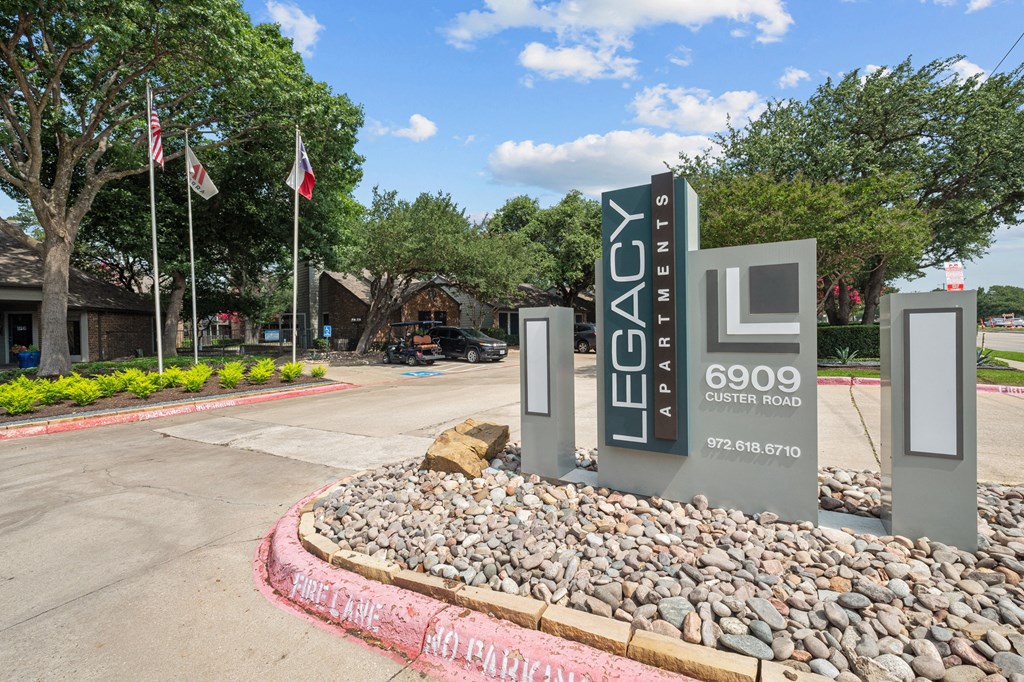 a sign that says Legacy Apartments at the entrance to a parking lot with flags in the background