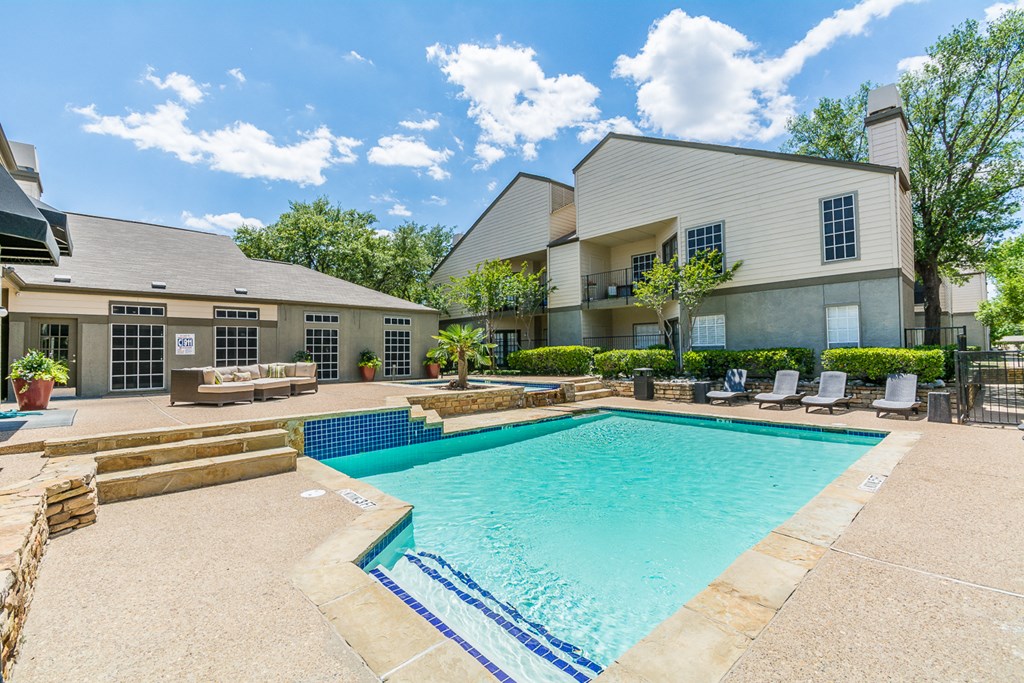 A swimming pool surrounded by a patio area with chairs and a building in the background.