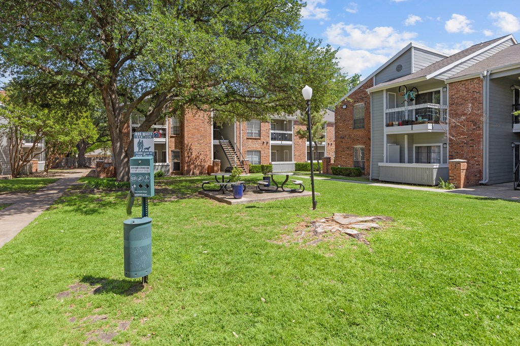 A green trash can is in the foreground of a grassy area in front of apartment buildings.