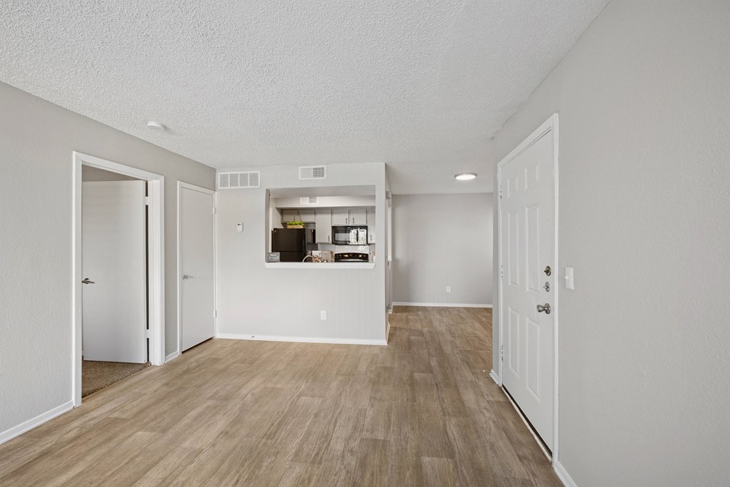 A room with white walls and wooden flooring with a kitchen visible through a doorway.