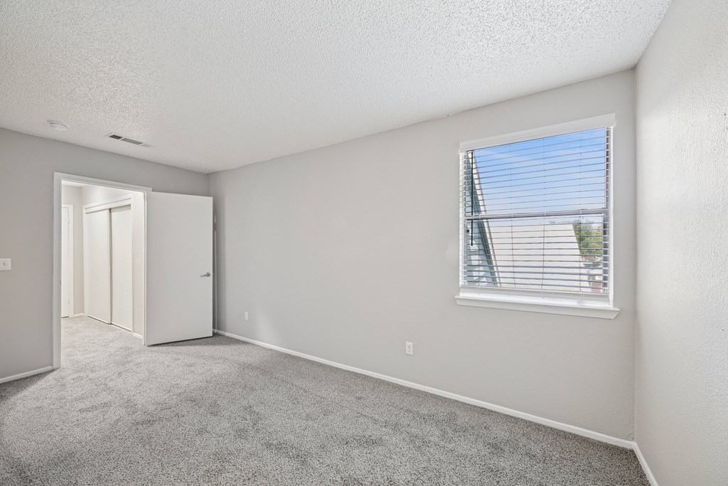 A room with a carpeted floor, a window with blinds, and a door leading to another room.