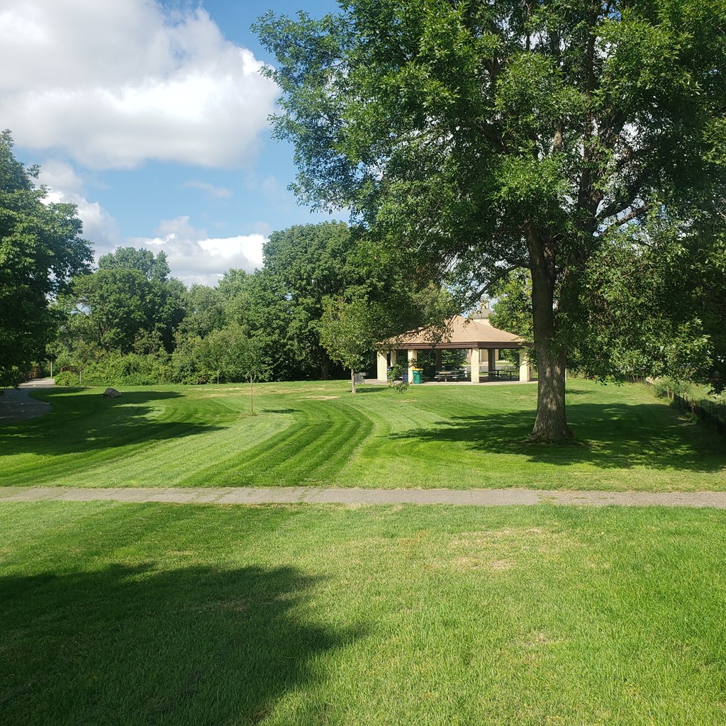 Large green pasture with a sidewalk across the middle. Gazebo with tables and chairs can be seen by the trees surrounding the grass. Blue and cloudy sky.