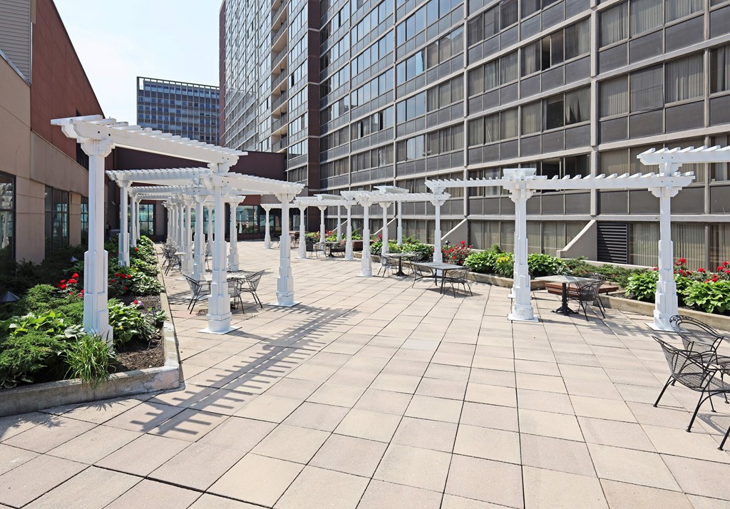A patio with white pergolas and chairs is surrounded by buildings.