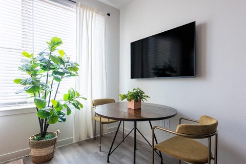 A living room with a round table, two chairs, a potted plant, and a flat screen TV mounted on the wall.