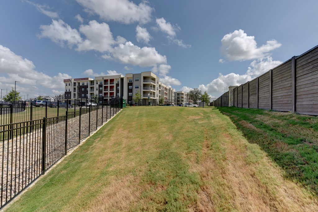 A long grassy area with a fence and apartment buildings in the background.