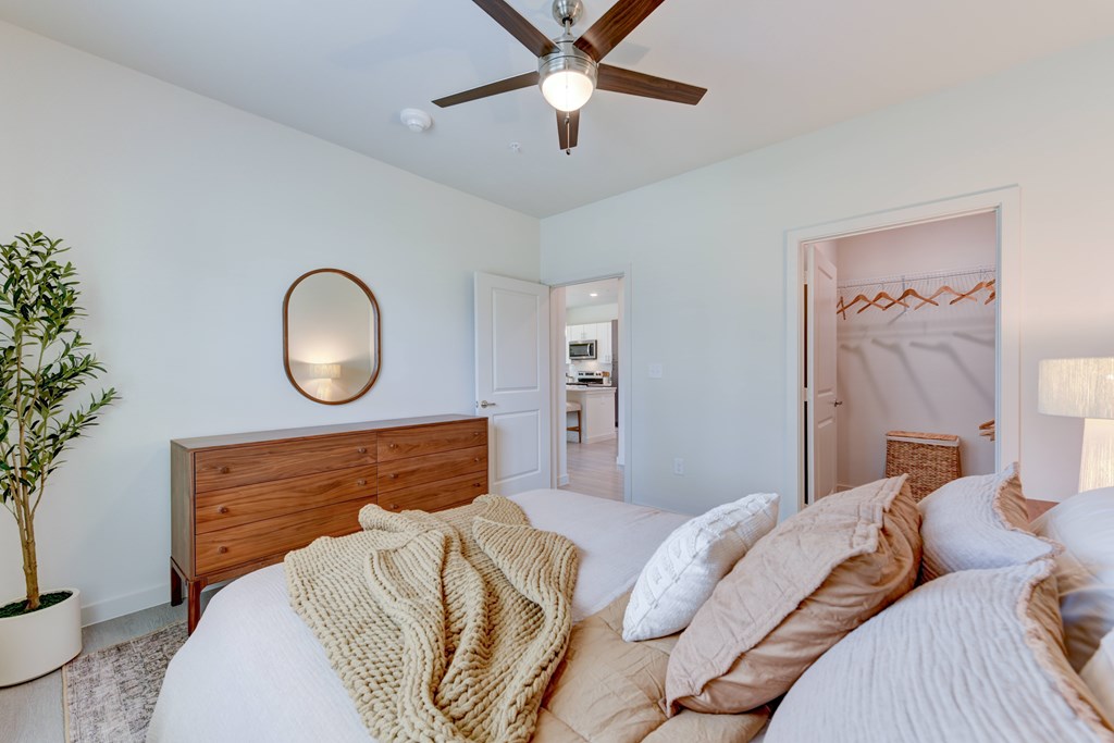 A bedroom with a bed covered in white and beige bedding, a ceiling fan, and a mirror on the wall.