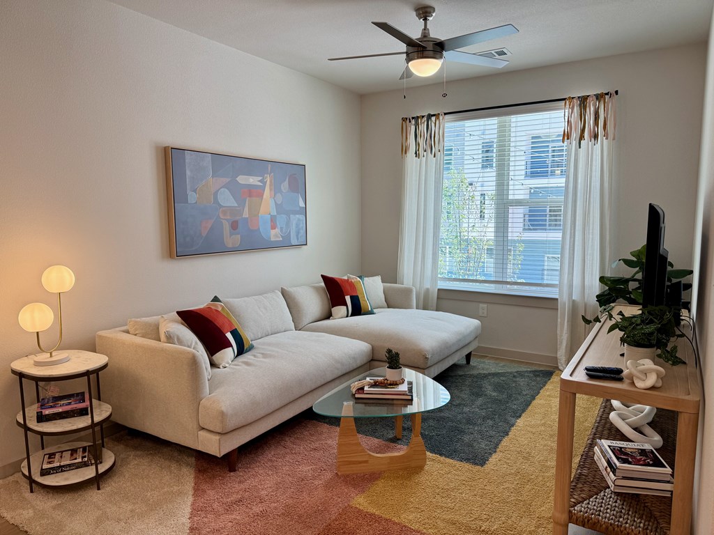 A living room with a white couch, a painting on the wall, a ceiling fan, and a window with curtains.
