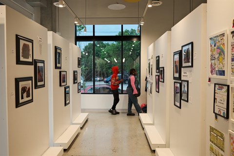 Two people are walking through an art gallery with white walls and pictures on the walls.
