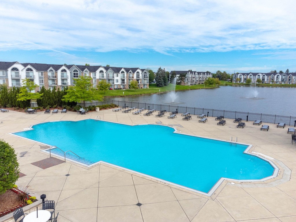 a large swimming pool with chaise lounge chairs next to a lake with apartment buildings in theat The Harbours Apartments, Clinton Twp, MI