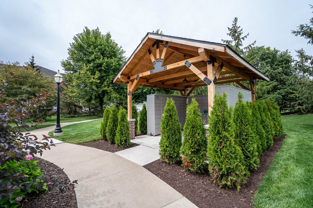 A wooden pavilion is surrounded by green shrubs and a concrete walkway.