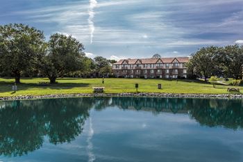 A serene landscape with a building, trees, and a calm lake. at Autumn Woods Apartments, Miamisburg, OH