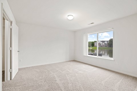 A room with a carpeted floor and a window showing a view of a house and trees at Towne Lakes Apartments, Grand Chute