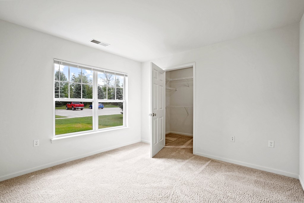 A room with a carpeted floor, a window with a view of a street, and a door leading to another room.