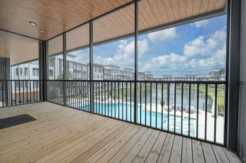 A wooden deck with a glass railing overlooking a pool and buildings.