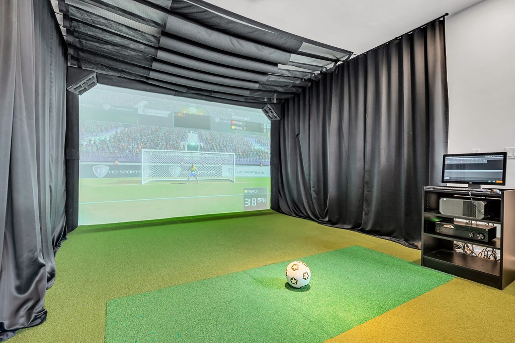 A soccer ball is on a green mat in front of a screen showing a soccer game at Luxe 360 on Centerpointe Apartments, Virginia, 23114