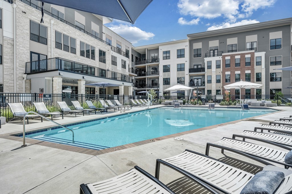 A swimming pool surrounded by sun loungers and umbrellas in front of apartment buildings.