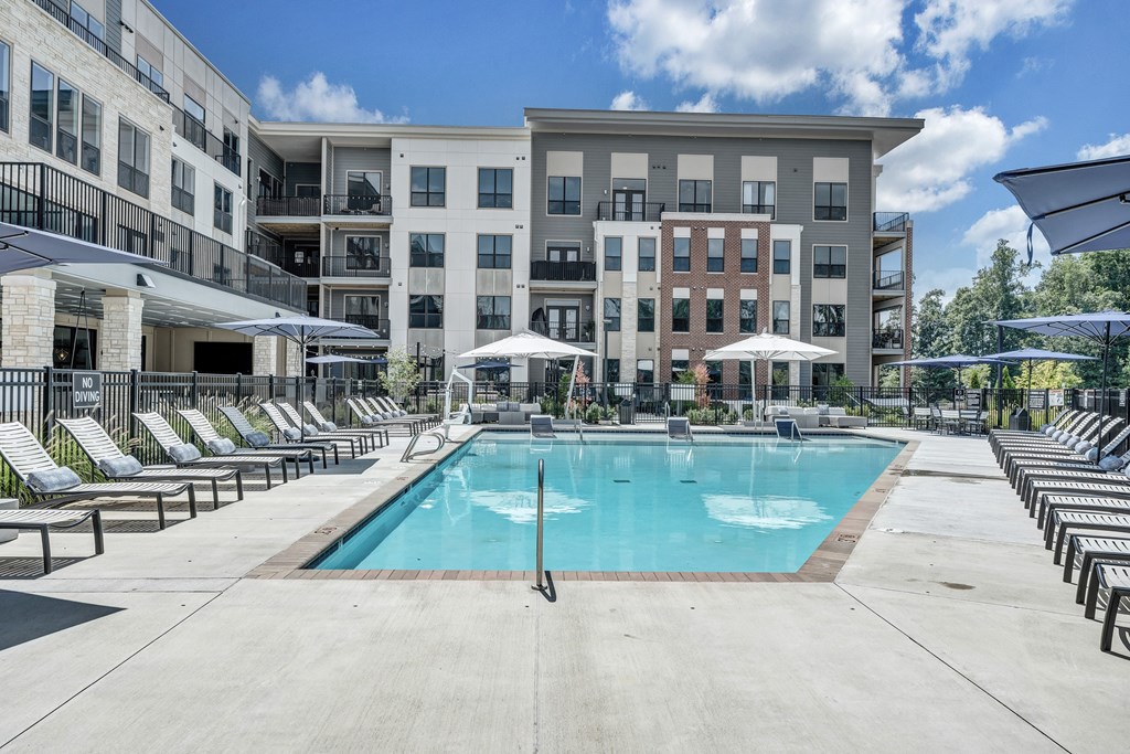 A large swimming pool surrounded by sun loungers and umbrellas in front of apartment buildings.