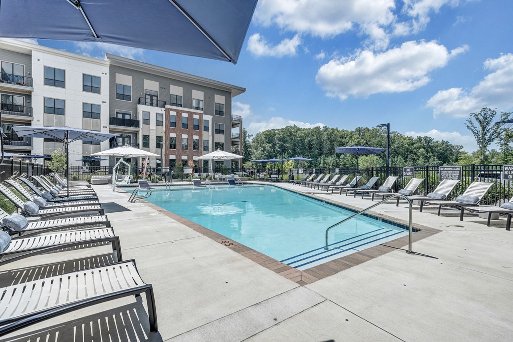 A swimming pool surrounded by sun loungers and umbrellas in front of apartment buildings.