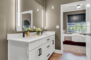 A bathroom with a vanity, mirror, and a bedroom in the background at Luxe 360 on Centerpointe Apartments, Virginia