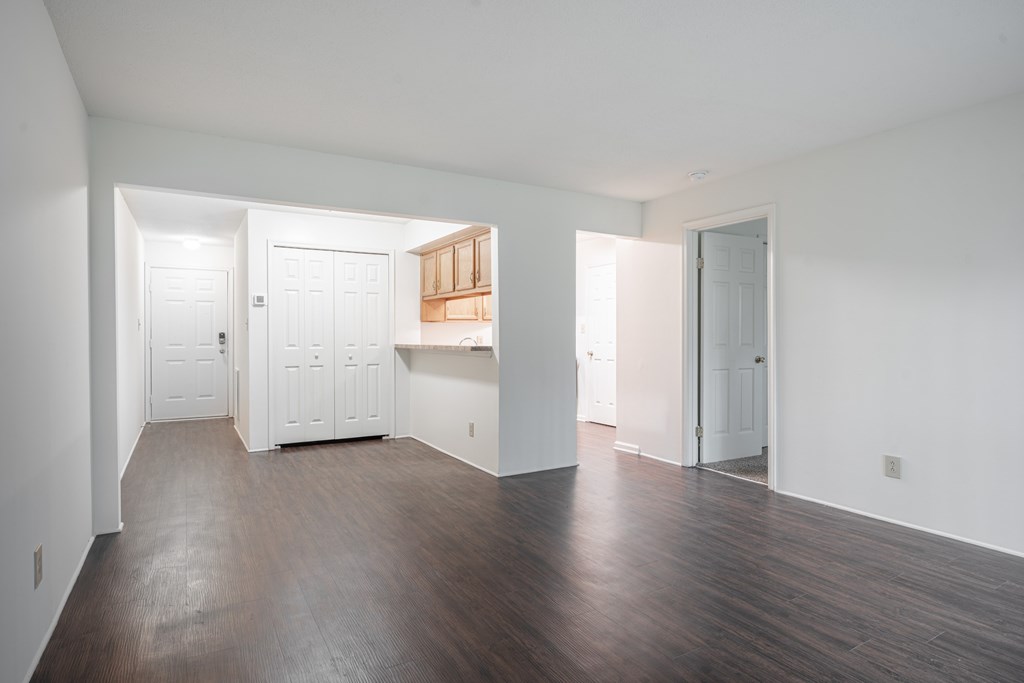 A room with white walls and wooden floors with a kitchen area in the middle.
