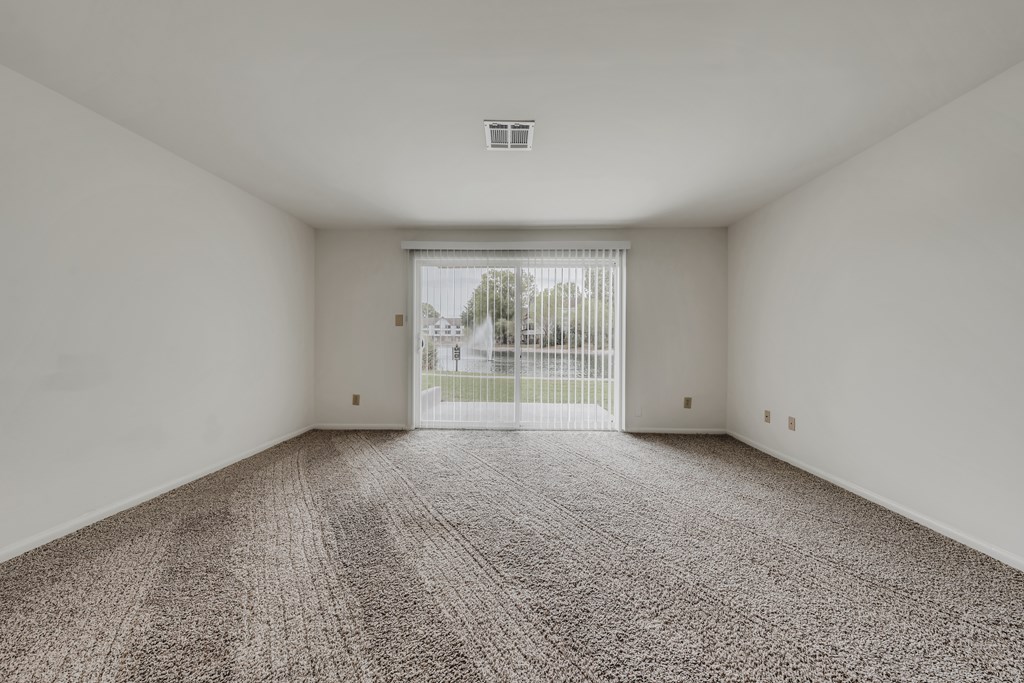 A room with a carpeted floor and a view of a fence and trees outside through a sliding glass door at WaterFront Apartments, Virginia Beach, 23453
