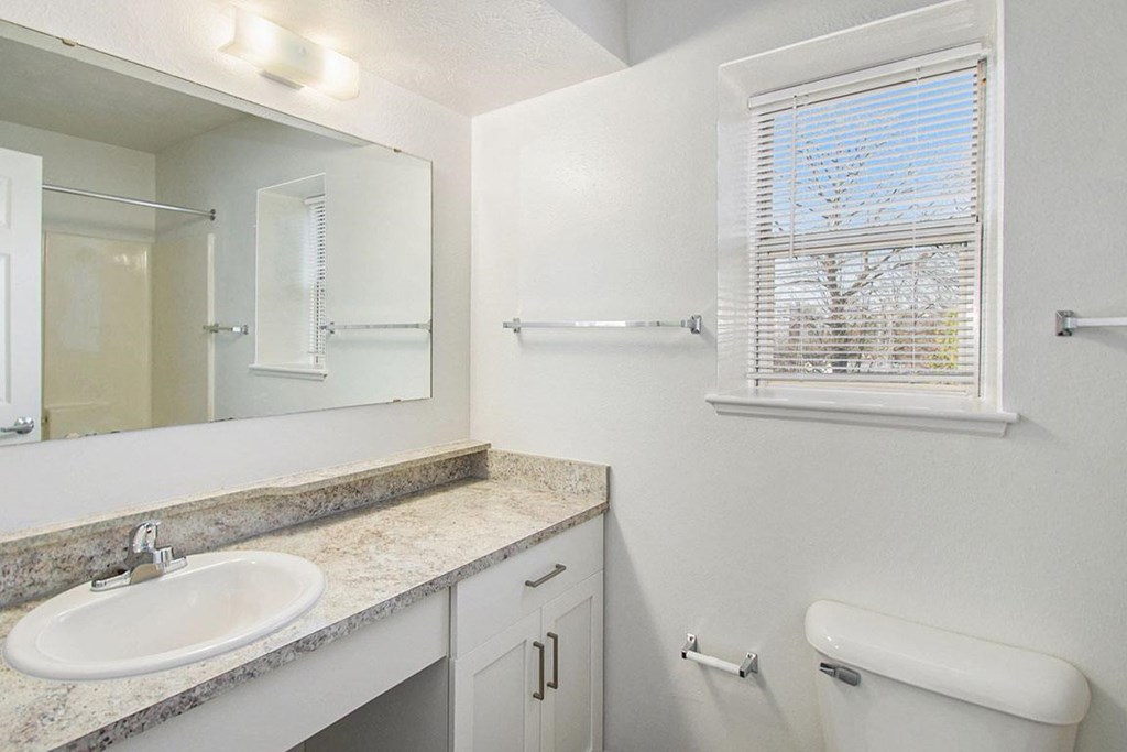 A bathroom with a sink, mirror, toilet, and a window with blinds at Brentwood Park Apartments, La Vista, Nebraska