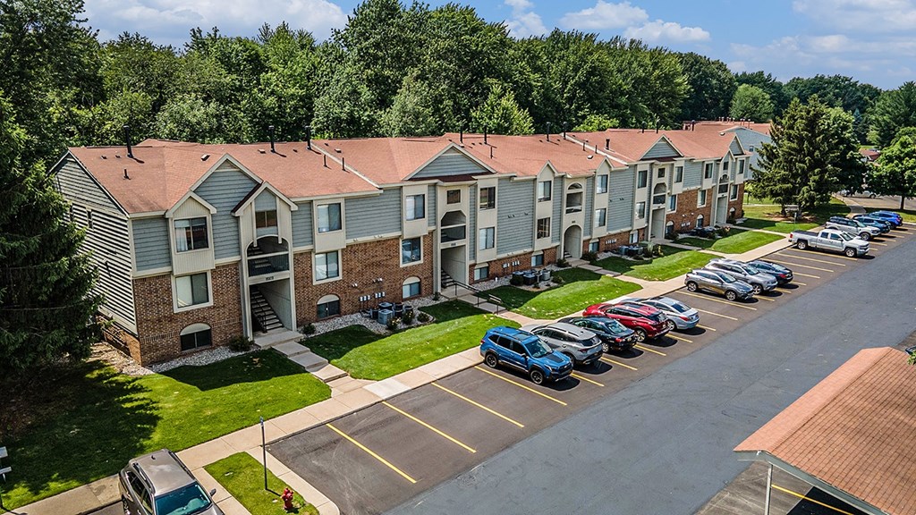 A parking lot in front of an apartment building with tall trees at Byron Lakes Apartments, Byron Center, Michigan