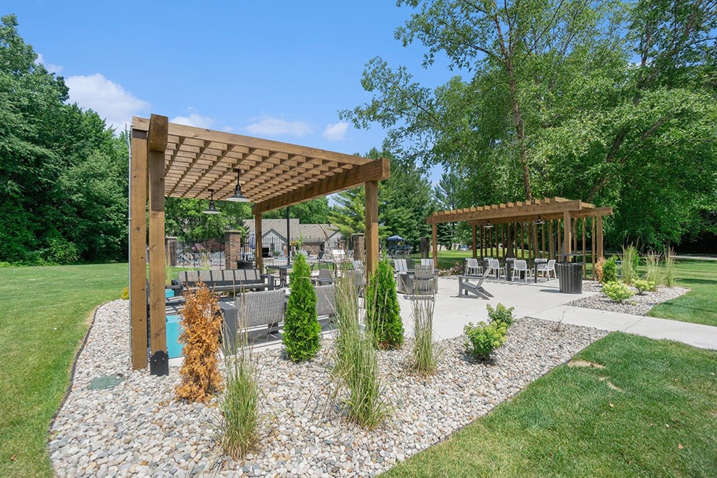 Wooden pergolas in the middle of a landscaped area with seating, a fire pit, and grills at Byron Lakes Apartments in Byron Center, MI