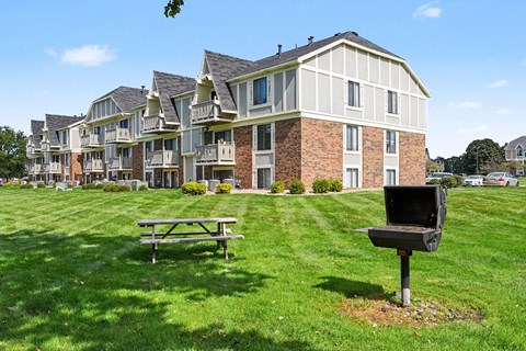 A bbq grill and picnic table in the foreground of a grassy area in front of apartment buildings at Normandy Village Apartments in Michigan City, IN