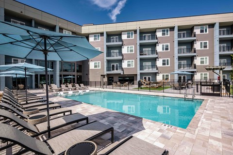 A large swimming pool surrounded by lounge chairs and umbrellas in front of apartment buildings.