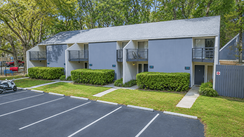 A parking lot in front of a grey building with trees in the background.