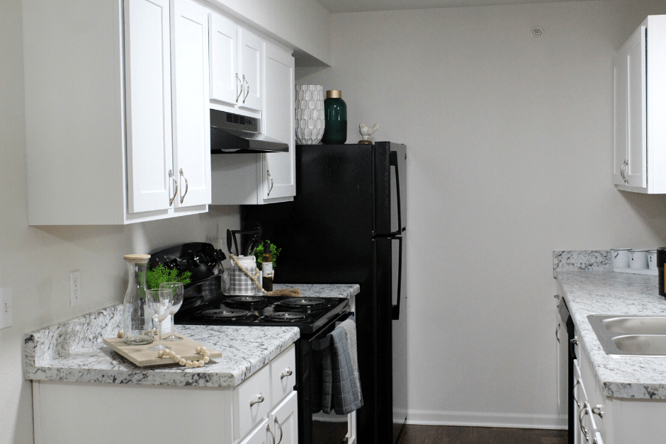 Updated kitchen at Riverwood. The walls are white with white cabinets on top and on bottom. There is a light speckled countertop with updated appliances. There is a stove to the left with a fridge and a sink and dishwasher to the right. There are dark wood-like flooring.