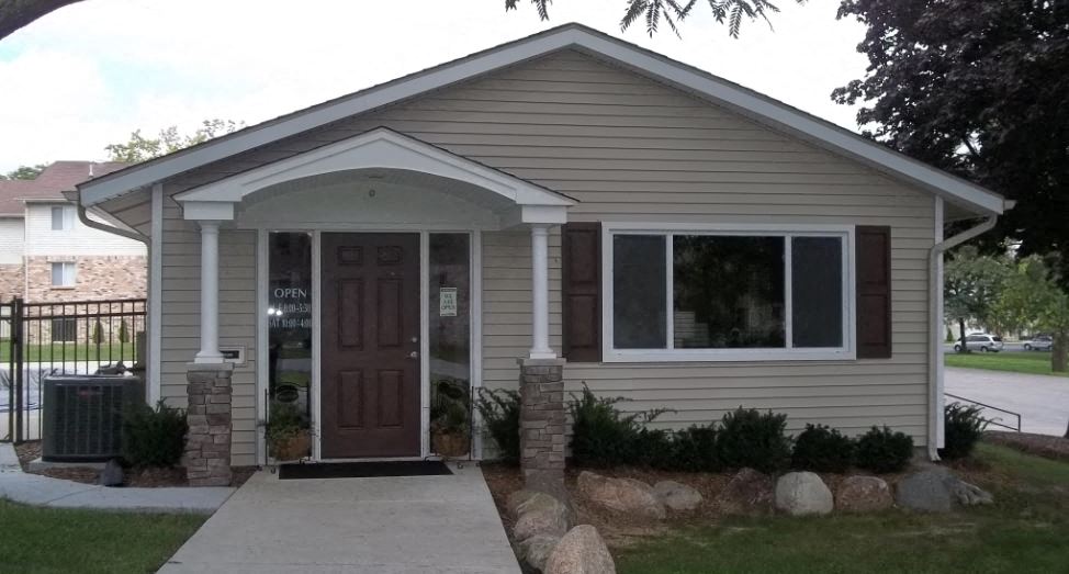 Tan building iwht white windows, brown doors, brown shutters, with brick pillars and landsacping with bushes nad rocks in front of buidling.  There is a large sidewalk leading up to the building. Behing the building to the left is an iron gate. In the background there is a brown brick/ brown sided building.