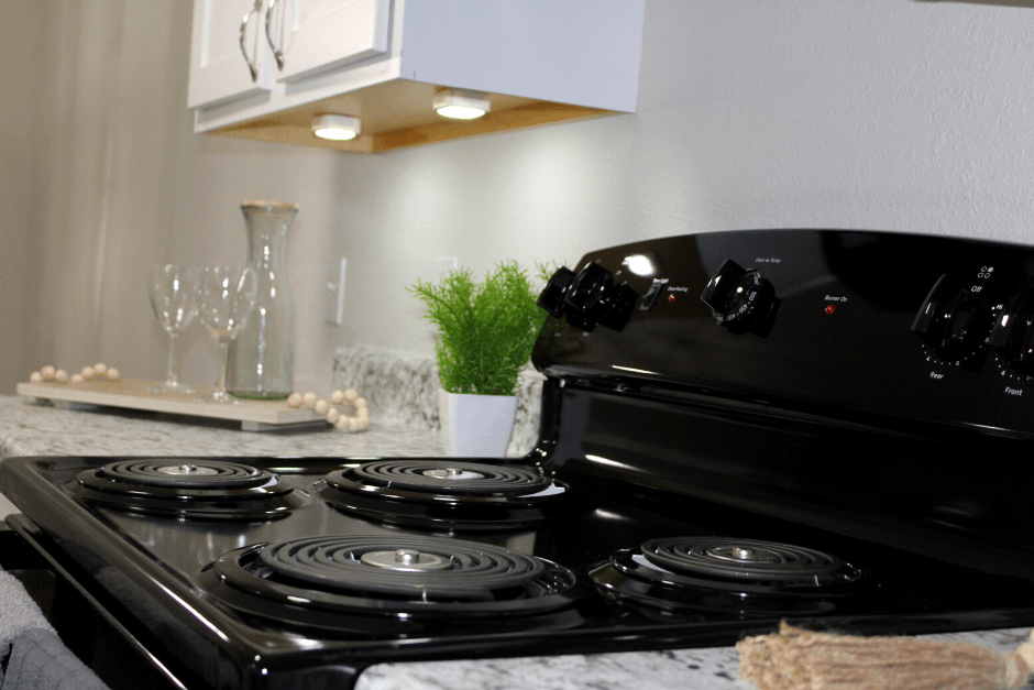 Photo of updated kitchen. There are white cabinets with a updated stove. There is a light specikled countertop. There is a green plant on the counter. There are lights underneath the cabinets.
