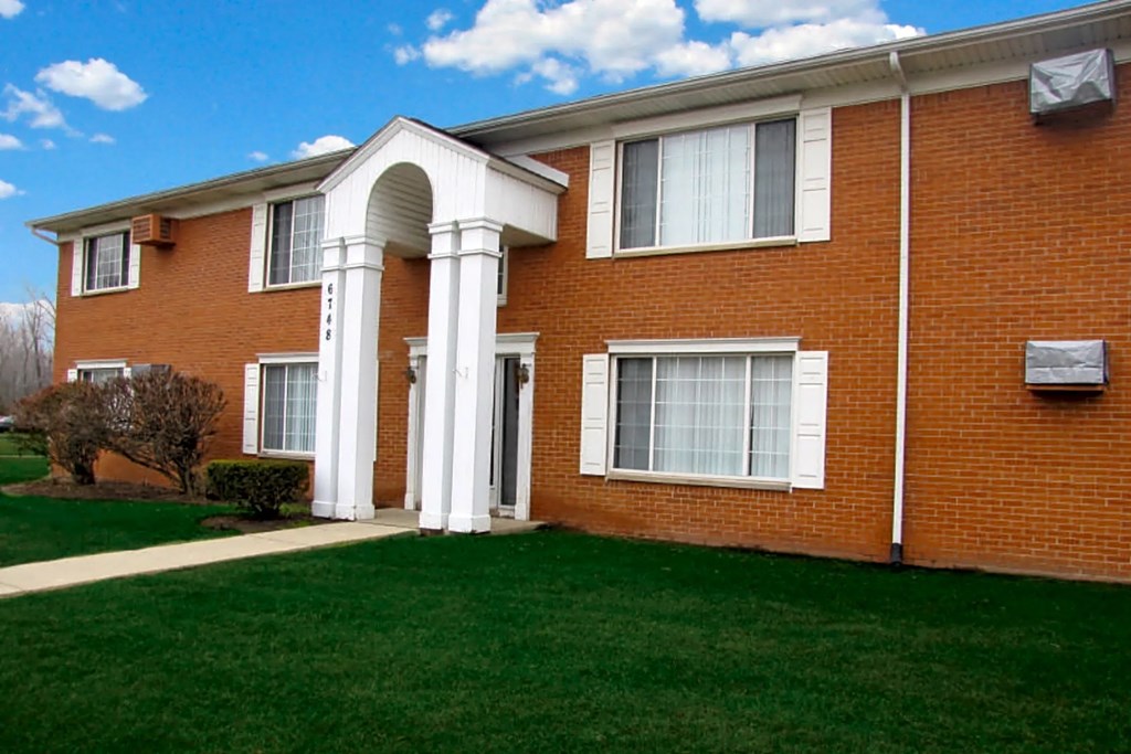 Red brick apartment building with tall white pillar entrance with green maintained lawn and blue sky.