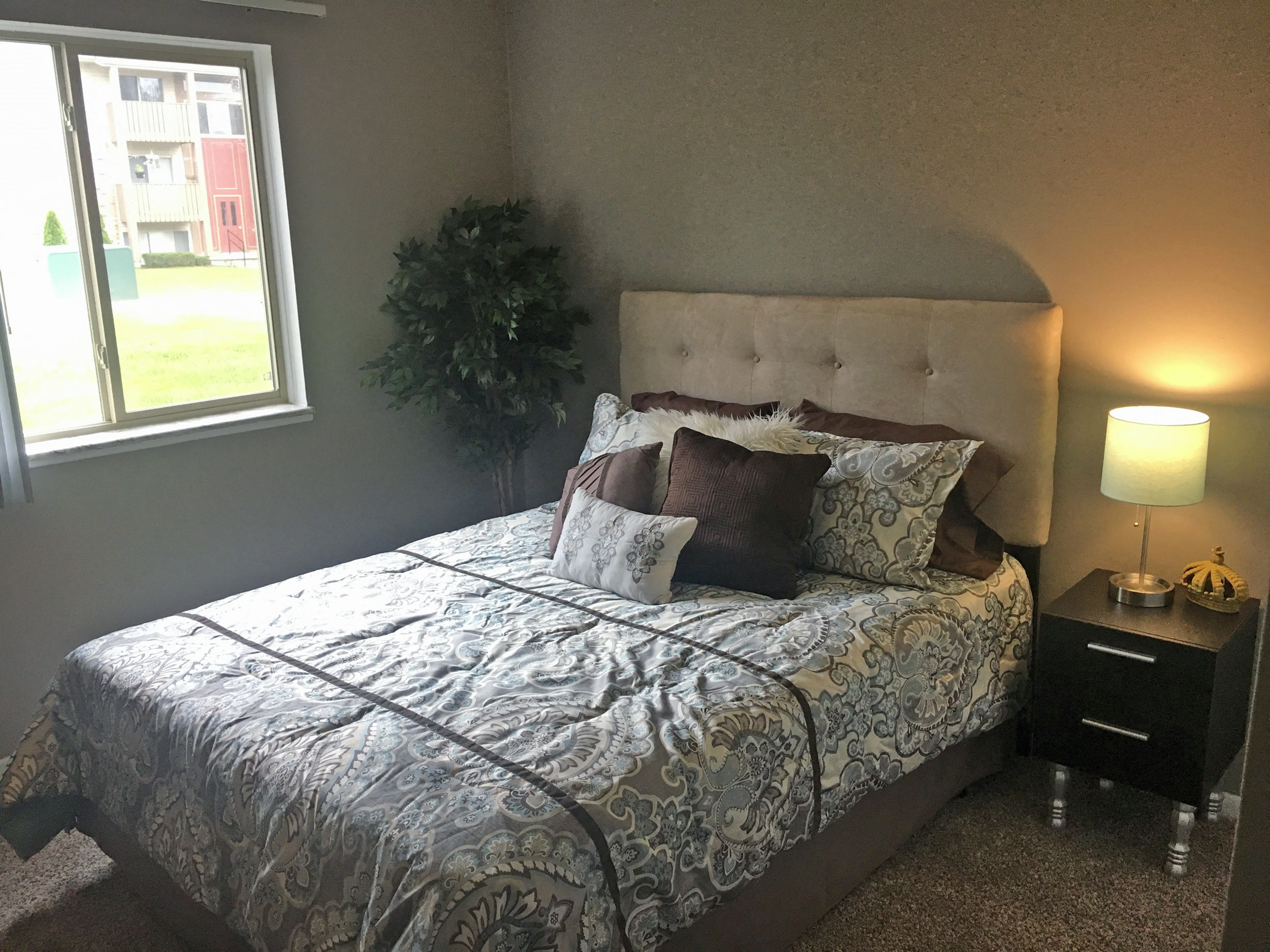 Bedroom with tan walls and tan carpet. The bed is made with a blue patterned bedding and brow and white accent pillows. There is a tan tufted headboard behind the pillows. A plant is in the left corner of the bedroom and a window takes up most of the left wall.
