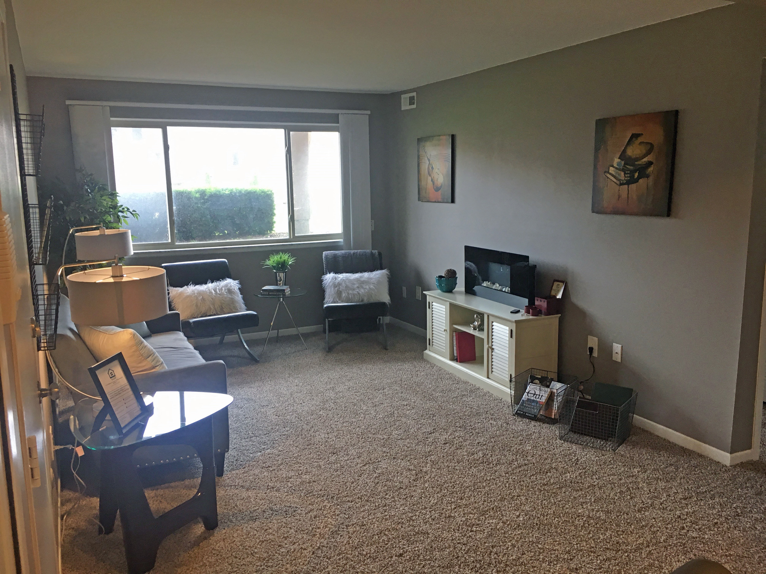 Living room with tan carpet and grey walls. There is a large pictured window on the back wall with two chairs under it. There is a couch against the left wall with a lamp and table next to it. Across from the couch on the right wall is a entertainment center. Art is also hanging on the right wall.