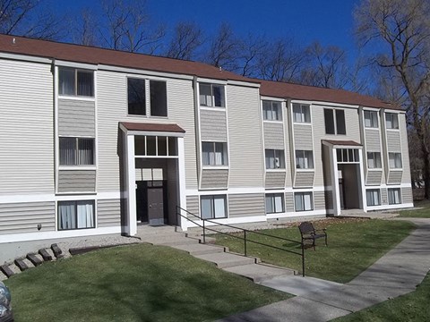 Exterior of building from the parking lot.  Sidwalk runs horizontal to building and there are 2 entrance doors.  Each door has white pillars on each side.  Building has beige siding with white trim.  Grass is green.