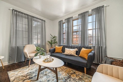 Image of a living room in an apartment at Sheridan Court. There are large windows on each side of the room. The walls are grey, with dark hardwood floors. There are two lounge chairs along with a dark navy couch. There is a patterned rug with a coffee table in the center of the room.