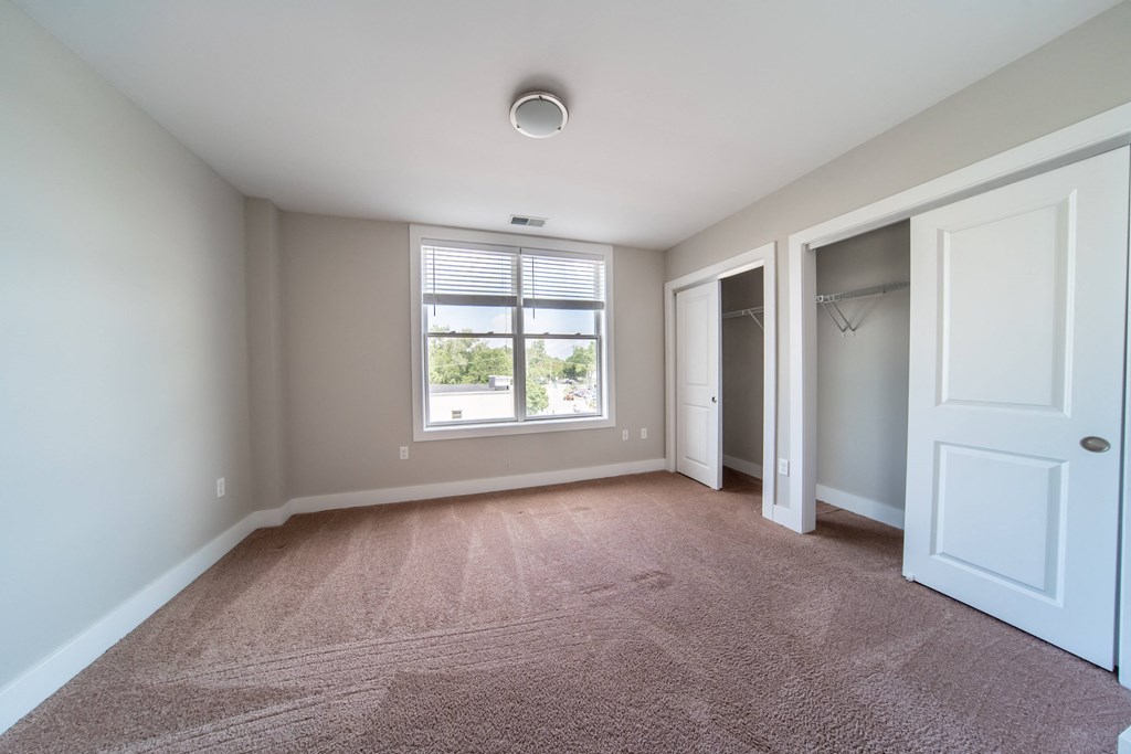 Bedroom with large window, beige plush carpeting, double closets, and tan walls.