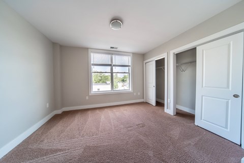 Bedroom with large window, beige plush carpeting, double closets, and tan walls.