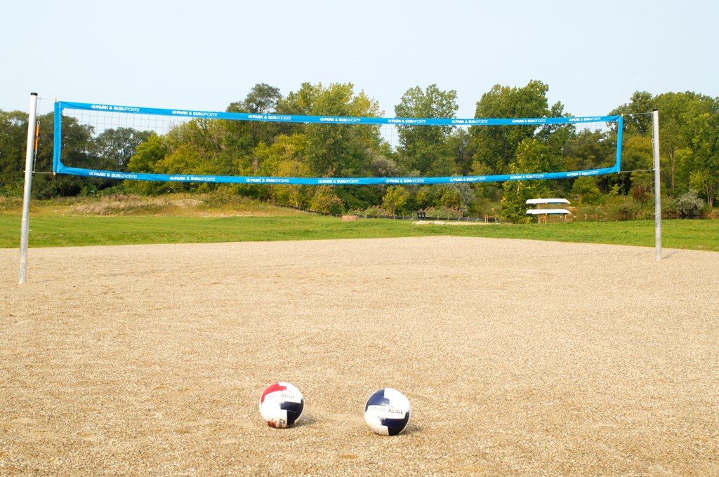 Outdoor sand volleyball court with a blue net. There are tow volleyballs on the court. Behind the court is a large grassy field and woods.