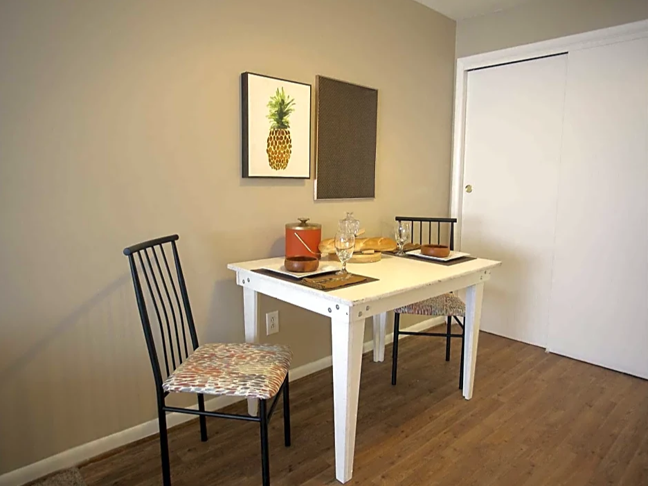 Dining Area with white table and 2 cloth covered rod iron chairs.  Pineapple photo above table on dark wood floor.  White doors behind table on right wall.